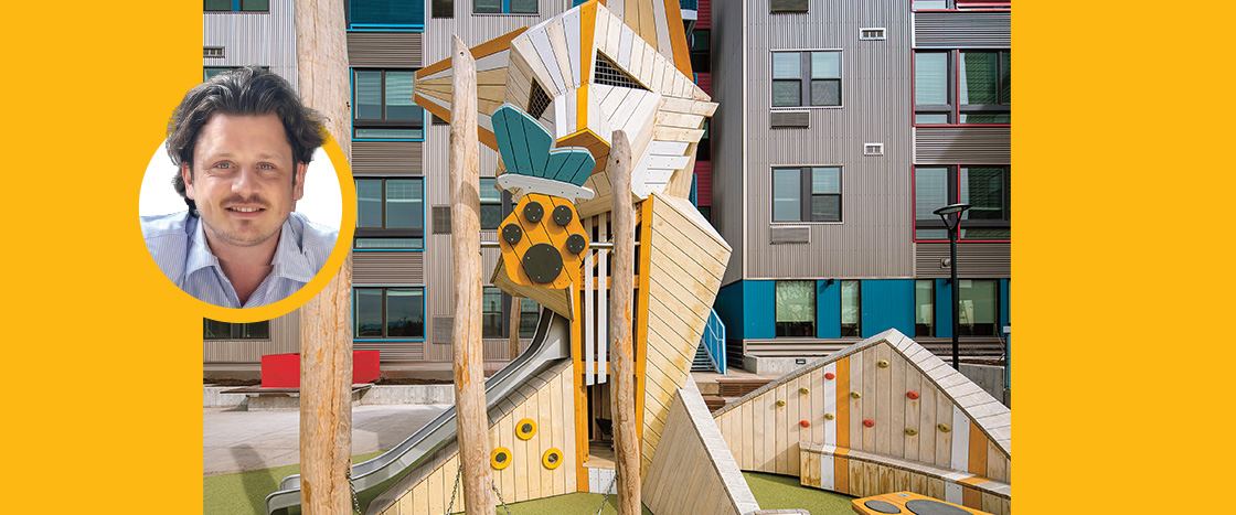 A man's portrait beside a tall wooden playground structure in an urban courtyard